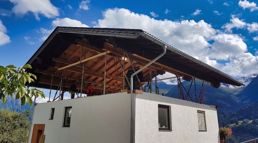Ein komplettes Satteldach schwebt auf hydraulischen Stützen deutlich über dem gemauerten Erdgeschoss eines weißen Hauses; in der Lücke sind Bauarbeiter und Stützkonstruktionen vor einer alpinen Bergkulisse zu sehen.
