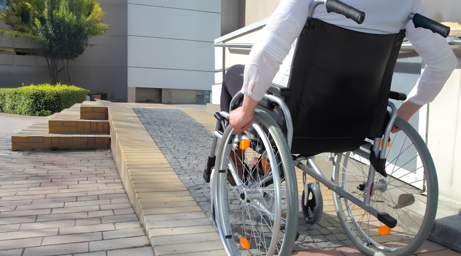 Eine Person in einem Rollstuhl fährt eine gepflasterte Rampe hinauf. Die Umgebung besteht aus modernen Gebäuden und grünen Pflanzen.