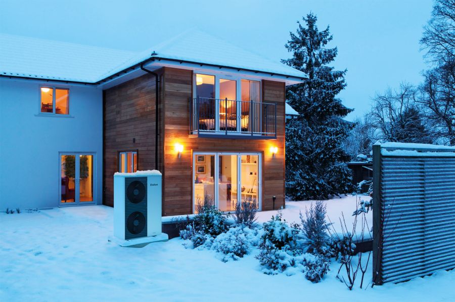 Eine Luft-Wasser-Wärmepumpe steht im verschneiten Außenbereich vor einem beleuchteten, modernen Einfamilienhaus in der Abenddämmerung. Die Außeneinheit ist teilweise mit Schnee bedeckt, während im Hintergrund warme Lichter aus den Fenstern des Hauses sche