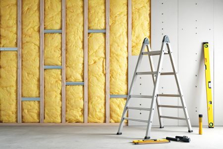 Das Bild zeigt eine im Bau befindliche Wand mit sichtbarer gelber Dämmung und der Rahmenkonstruktion. Eine Leiter steht davor, und Werkzeuge liegen auf dem Betonboden, was auf Bauarbeiten hindeutet. Die Szene vermittelt eine Atmosphäre des Fortschritts un