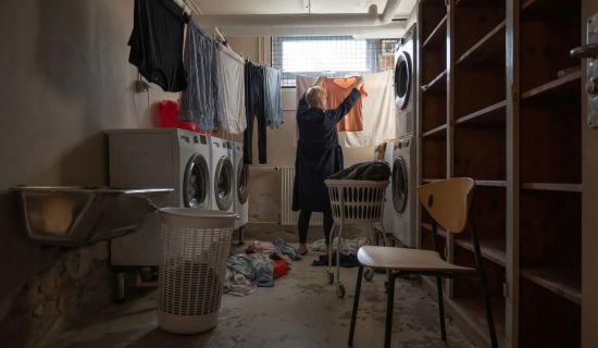 Ein funktionaler Kellerraum, der als Waschküche dient, mit mehreren weißen Waschmaschinen an der Wand. Eine Person steht mit dem Rücken zur Kamera und hängt ein orangefarbenes Kleidungsstück auf eine Wäscheleine, an der bereits andere Kleidung trocknet.