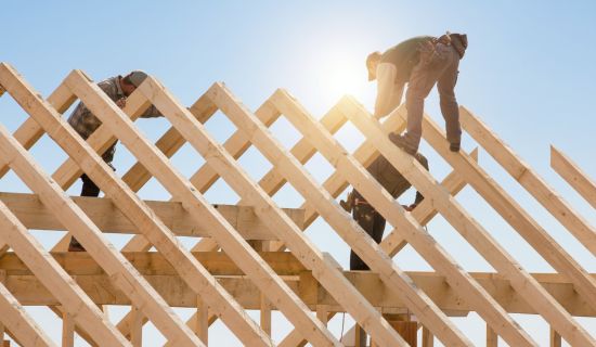 Handwerker in Arbeitskleidung balancieren auf den hölzernen Balken eines offenen Dachstuhls unter strahlend blauem Himmel und montieren die Konstruktion.