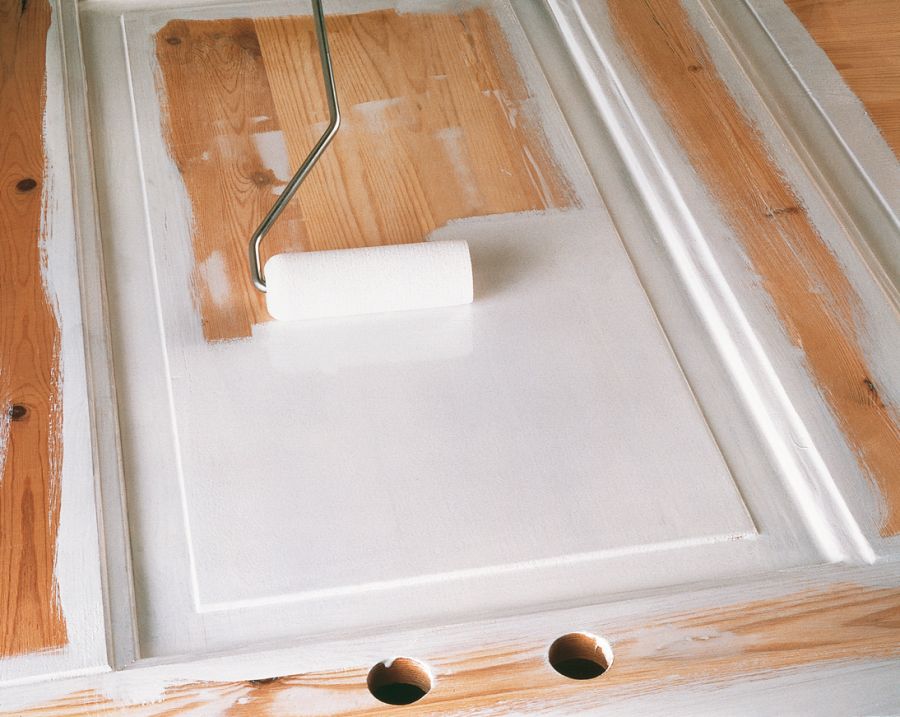 Eine weiße Lackrolle wird über die Vertiefung einer holzfarbenen Kassettentür geführt. Die innere Fläche der Kassette ist bereits weiß lackiert, während der Rahmen der Tür teilweise grundiert und teilweise noch blankes Holz ist.