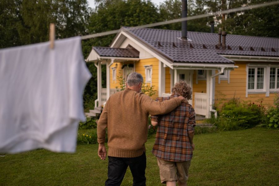 Altes Paar geht auf ein altes Haus zu, der Mann stützt die Frau
