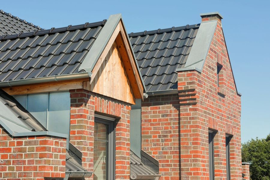 Außenansicht eines modernen Einfamilienhauses mit einer rot-blau-bunten Klinkerfassade und anthrazitfarbenem Dach. Die Detailaufnahme zeigt Giebel, Dachüberstände und in das Mauerwerk eingelassene Fenster bei sonnigem Wetter.