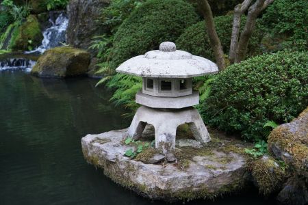 Eine traditionelle japanische Steinlaterne steht auf einem moosbewachsenen Felsen am Rande eines ruhigen Teichs. Im Hintergrund sind grüne Pflanzen und ein kleiner Wasserfall zu erkennen, die eine meditative Atmosphäre schaffen.