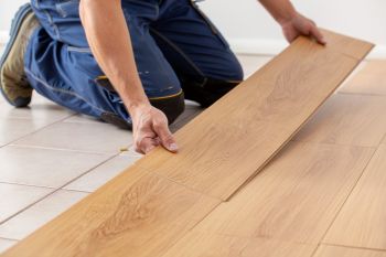 Das Bild zeigt einen Handwerker in blauer Arbeitskleidung, der auf hellen Fliesen kniet und einen neuen hellen Laminatboden verlegt. Er ist dabei, eine weitere Holzdiele präzise in den bereits verlegten Bereich einzufügen, was die Szene von konzentrierter