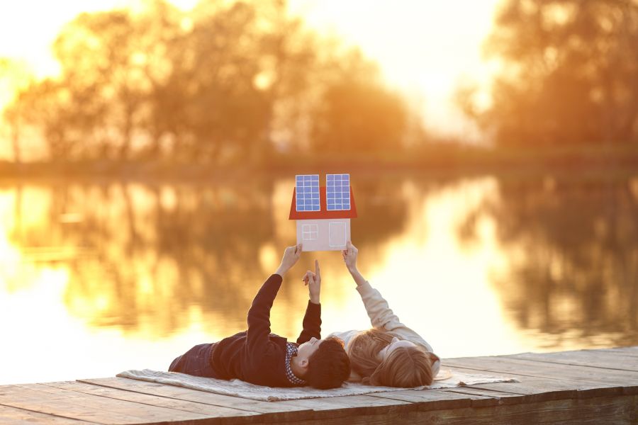 Zwei Kinder liegen auf einem Steg am See und hält ein kleines Hausmodell mit Solarzellen in die Höhe, was den Traum vom energieautarken Eigenheim symbolisiert.