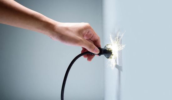 Eine Hand steckt ein schwarzes Stromkabel in eine weiße Wandsteckdose. Dabei entsteht ein heller, sprühender Funkenflug direkt am Kontakt zwischen dem Stecker und der Dose. Die Wand im Hintergrund ist schlicht und in einem hellen Grauton gehalten.