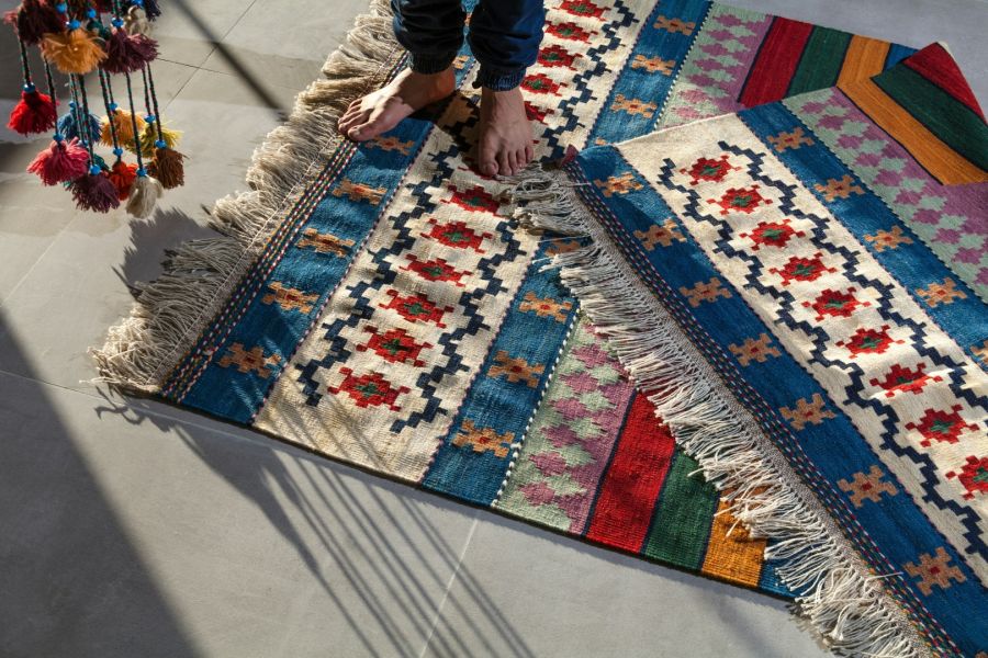 Eine Person steht barfuß auf einem flachgewebten Kelim-Teppich, der komplexe geometrische Muster in Blau, Rot und Grün aufweist. Der Teppich ist an einer Ecke umgeklappt und zeigt lange, helle Fransen, während im linken Bildrand bunte Quasten im Sonnenlic
