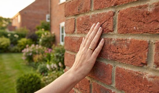 Eine weibliche Hand mit einem goldenen Ring berührt flach eine rote Backsteinfassade. Der Hintergrund zeigt unscharf einen gepflegten Garten und Nachbarhäuser im Abendlicht.