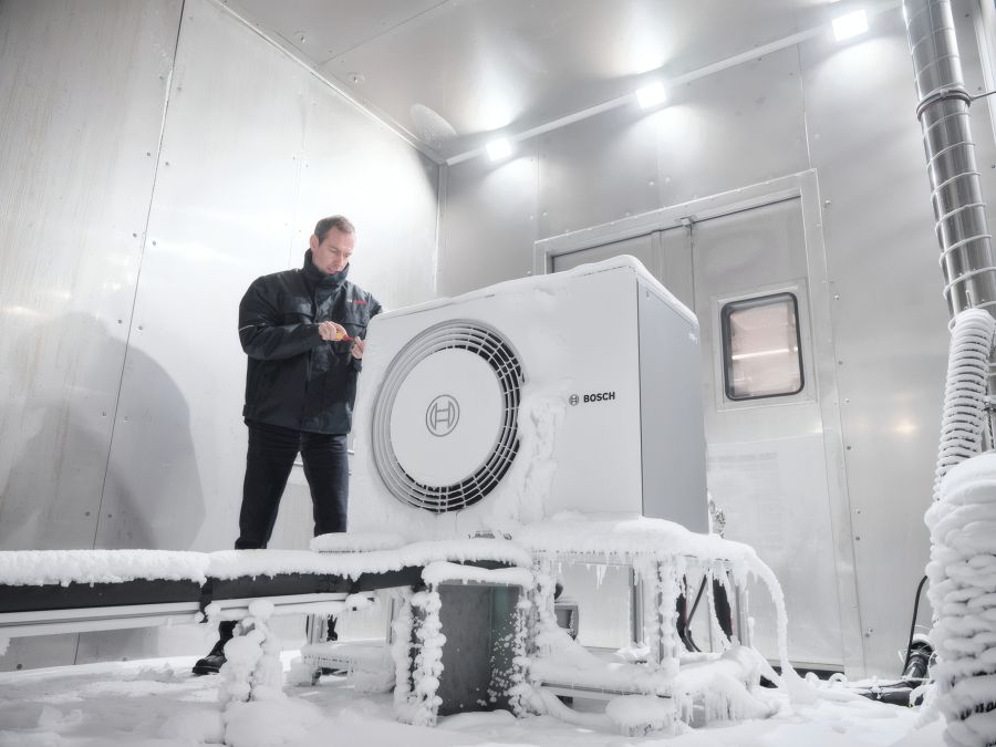 Ein Techniker in Winterkleidung prüft eine stark vereiste Wärmepumpe in einem silbernen Laborraum. Das Gerät und die umliegenden Leitungen sind mit einer dicken Schicht aus Frost und Eiszapfen bedeckt.