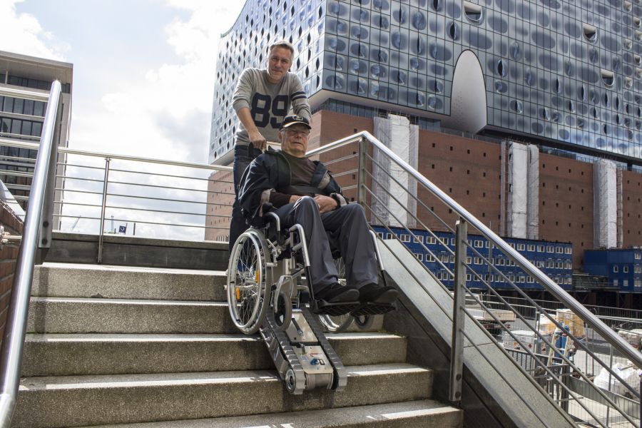Mann schiebt älteren Mann im Rollstuhl mit Treppenraupe eine Betontreppe in urbaner Umgebung hinunter, im Hintergrund die Elbphilharmonie.