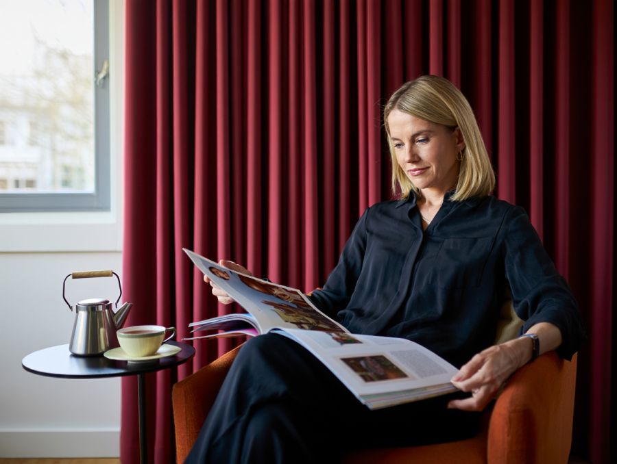Eine Frau sitzt in einem bequemen Sessel und liest entspannt ein Magazin. Im Hintergrund sind rote Vorhänge und ein Fenster zu sehen, daneben steht ein kleiner Tisch mit Teekanne und Tasse, was eine ruhige und gemütliche Atmosphäre schafft.
