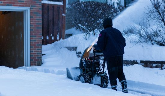 Eine Person in dunkler Winterkleidung schiebt eine motorisierte Schneefräse über einen verschneiten Gehweg oder eine Einfahrt. Hoher Schnee türmt sich an den Rändern auf, während die Maschine den Schnee zur Seite wirft.