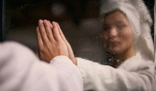 Eine Nahaufnahme von zwei Händen, die einen vollständig beschlagenen Spiegel berühren. Das Spiegelbild der Person im weißen Bademantel ist durch das Kondenswasser nur verschwommen erkennbar.