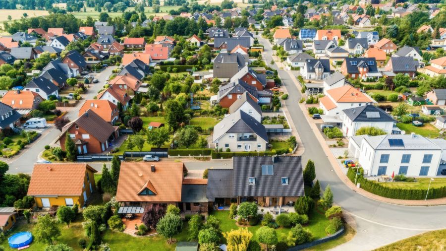 Eine Drohnenaufnahme zeigt eine weitläufige Vorstadtsiedlung mit zahlreichen freistehenden Einfamilienhäusern. Die Grundstücke sind mit grünen Gärten durchsetzt, und die Gebäude weisen überwiegend rote sowie dunkle Satteldächer auf. Das Bild veranschaulic