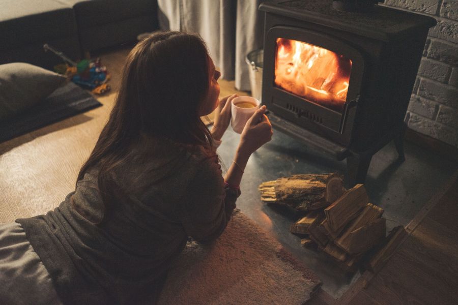Eine Frau liegt entspannt auf dem Boden und hält eine Tasse, während sie in das Feuer eines Kaminofens blickt, Holzscheite liegen daneben.