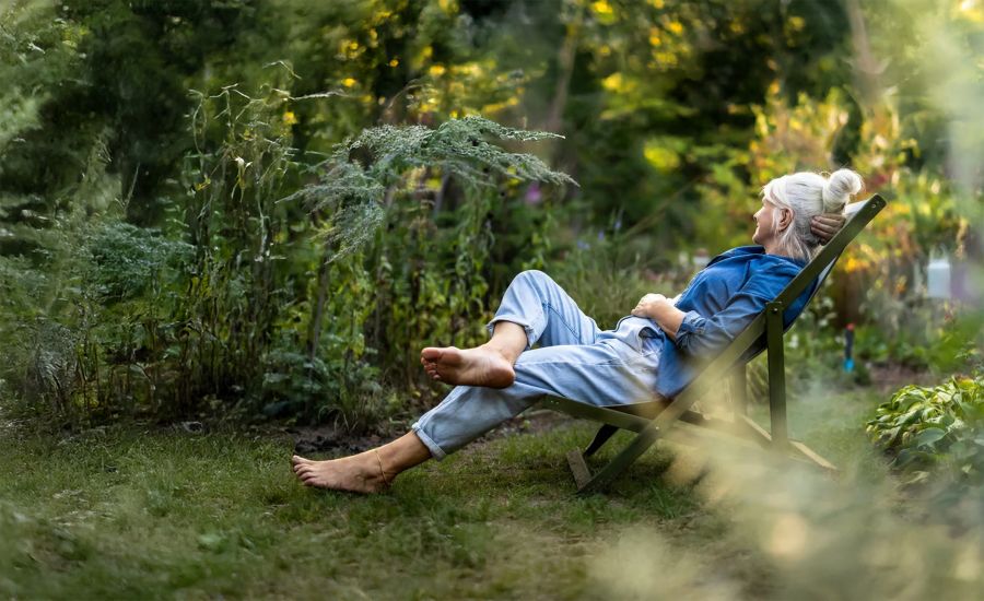 Eine Frau mit hochgesteckten grauen Haaren entspannt barfuß in einem Liegestuhl inmitten eines naturbelassenen, grünen Gartens. Das weiche Licht und die üppige Umgebung vermitteln ein Gefühl von Frieden und Erholung.