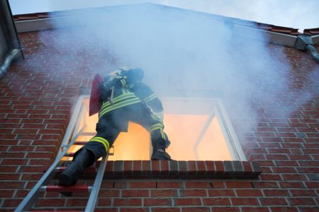 Ein Feuerwehrmann in voller Schutzausrüstung steigt über eine Leiter in das Fenster eines brennenden Backsteinhauses. Dichter grauer Rauch quillt aus dem Gebäude und steigt an der Fassade empor.