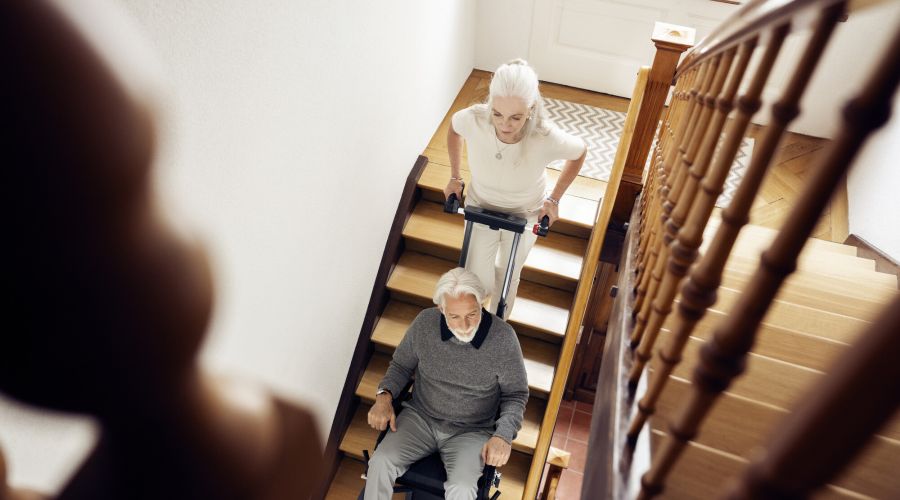 Ältere Frau fährt ihren Partner im Treppensteiger eine Holztreppe im Haus hinunter. Beide wirken entspannt.