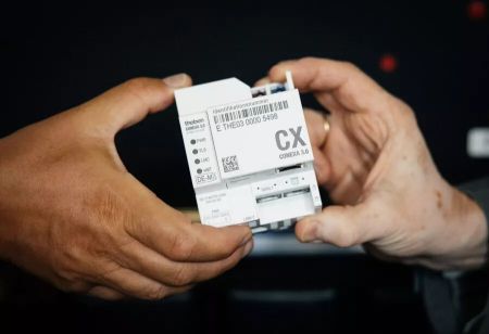 Zwei Hände halten ein kompaktes, weißes Smart-Meter-Gateway des Typs CONEXA 3.0 für den Einbau im Zählerschrank. Auf dem Elektronikgehäuse sind Identifikationsnummer, Barcodes, Anschlussbuchsen und Kontroll-LEDs gut lesbar abgebildet. Das Gerät wird front