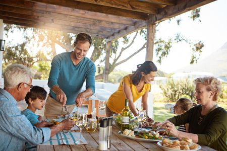 Eine fröhliche Familie sitzt an einem Holztisch unter einer überdachten Terrasse und genießt ein gemeinsames Essen. Ein Mann in einem blauen Pullover schneidet Fleisch, während eine Frau in einem gelben Oberteil lacht. Ältere und jüngere Familienmitgliede