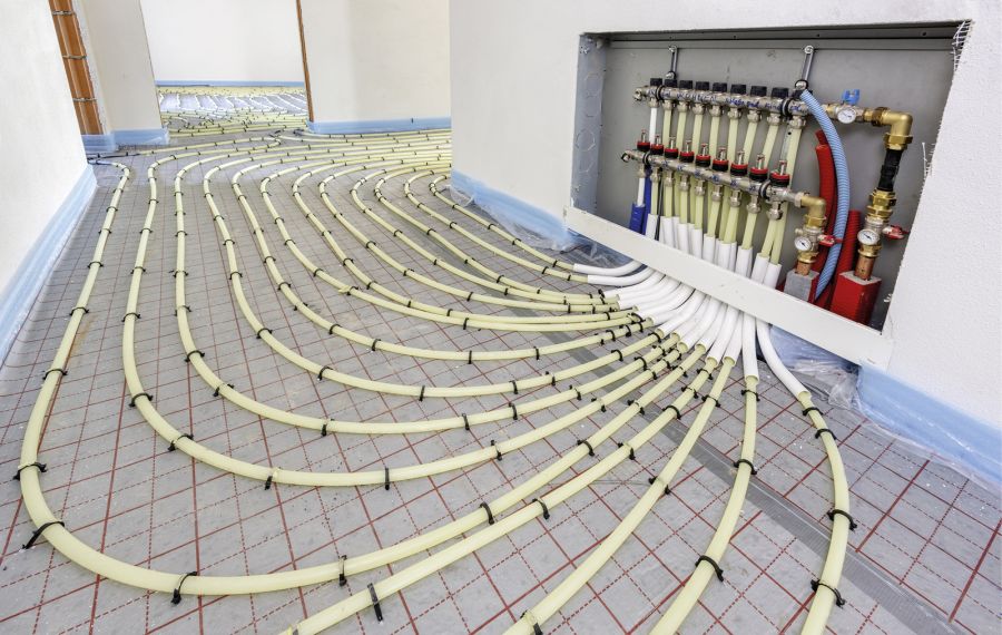 Das Bild zeigt eine Fußbodenheizung mit gelben Rohren, die in geschwungenen Linien auf einem Boden verlegt sind. Im Hintergrund ist eine Wand mit einem Heizkreisverteiler, an dem mehrere farbige Rohre und Ventile sichtbar sind.