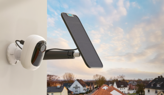 Eine moderne, weiße Sicherheitskamera ist zusammen mit einem kleinen Solarpanel an einer hellen Hauswand montiert. Im Hintergrund ist eine Wohnsiedlung bei dämmerndem Himmel mit sanften Wolkenformationen zu sehen.