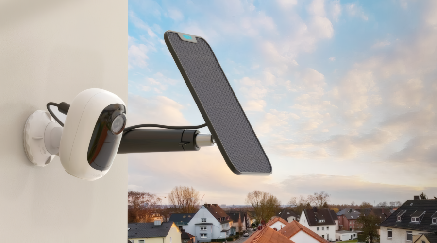Eine moderne, weiße Sicherheitskamera ist zusammen mit einem kleinen Solarpanel an einer hellen Hauswand montiert. Im Hintergrund ist eine Wohnsiedlung bei dämmerndem Himmel mit sanften Wolkenformationen zu sehen.