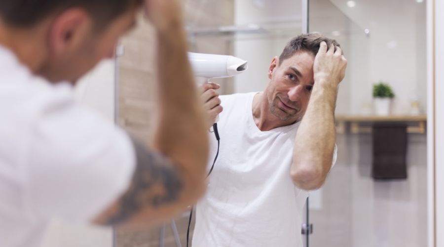 Ein Mann in einem weißen T-Shirt steht im Badezimmer vor dem Spiegel und föhnt sich die Haare. Er betrachtet sein Spiegelbild, das klar zu erkennen ist, was auf eine gute Belüftung hindeutet.