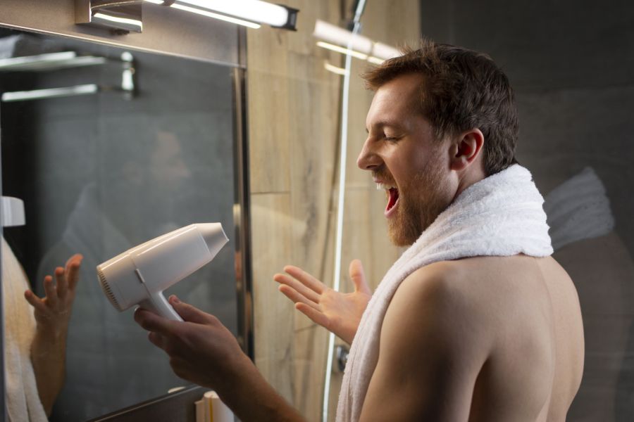 Ein Mann mit Bart und einem weißen Handtuch um die Schultern steht seitlich im Badezimmer. Er hält einen weißen Föhn wie ein Mikrofon und singt enthusiastisch hinein.