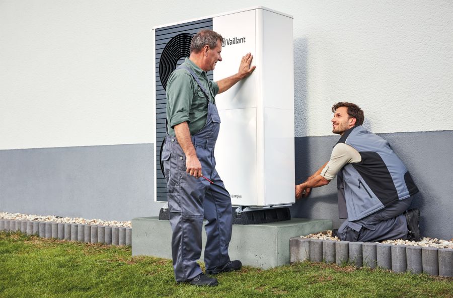Zwei Handwerker in grauer Arbeitskleidung montieren eine weiße Luft-Wasser-Wärmepumpe auf einem massiven Betonsockel an einer Hauswand. Sie arbeiten bei Tageslicht im Freien und richten das Gehäuse des Geräts präzise aus.