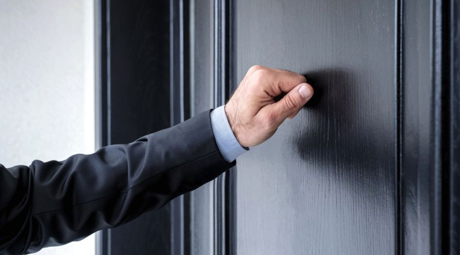 Eine männliche Hand im dunklen Anzug klopft an eine geschlossene, schwarz lackierte Holztür. Die Geste symbolisiert das Verlangen nach Einlass in eine Wohnung.