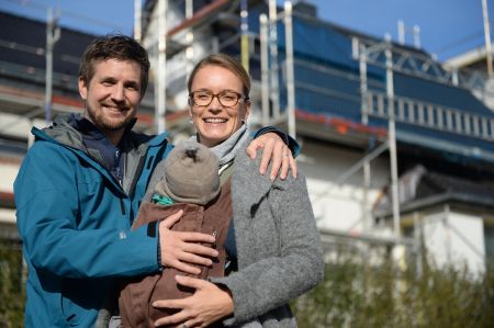 Eine dreiköpfige junge Familie steht lächelnd vor einem Wohnhaus, das für anstehende Sanierungsarbeiten komplett mit einem Baugerüst eingerüstet ist.