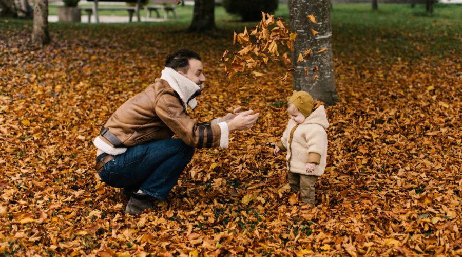 Ein Mann in einer braunen Jacke kniet im Freien auf einem Boden, der vollständig mit goldenen Herbstblättern bedeckt ist. Er spielt mit einem kleinen Kind in einer beigen Jacke, das vor ihm steht, und wirft Blätter in die Luft.