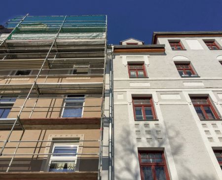 Zwei direkt aneinandergrenzende mehrstöckige Wohngebäude sind von unten aus der Froschperspektive fotografiert. Die linke Fassade ist komplett mit einem Baugerüst versehen und wird saniert, während die rechte, weiß gestrichene Fassade unsaniert im Bestand