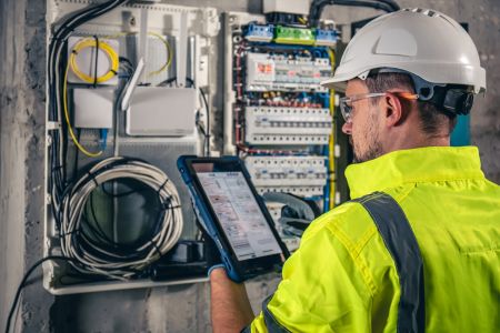 Ein Elektrotechniker mit weißem Schutzhelm, Schutzbrille und neongelber Warnweste steht vor einem geöffneten Sicherungskasten. Er hält ein blaues Tablet in der linken Hand und tippt mit der rechten auf den Bildschirm, während er die Installationen überprü