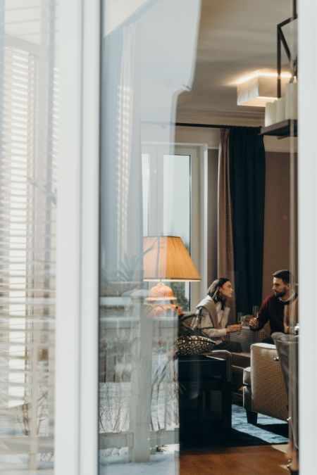Ein Paar sitzt entspannt mit Getränken in der Hand in einem behaglich beleuchteten Wohnzimmer. Die Szene wird von außen durch ein Fenster betrachtet, was eine warme, intime und einladende Atmosphäre schafft.