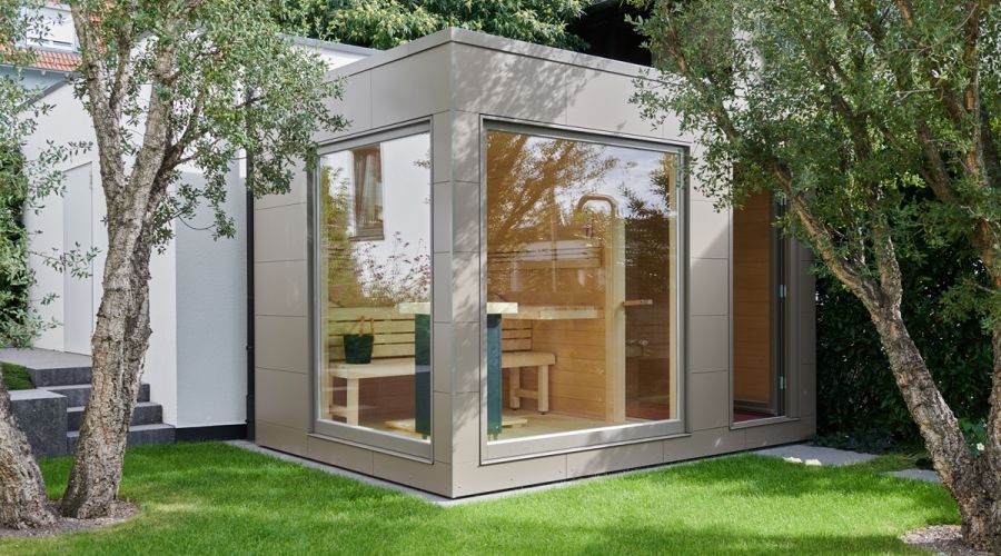 Moderne Gartensauna mit grauer Fassade, großen Panoramafenstern und Flachdach zwischen Bäumen im Garten.