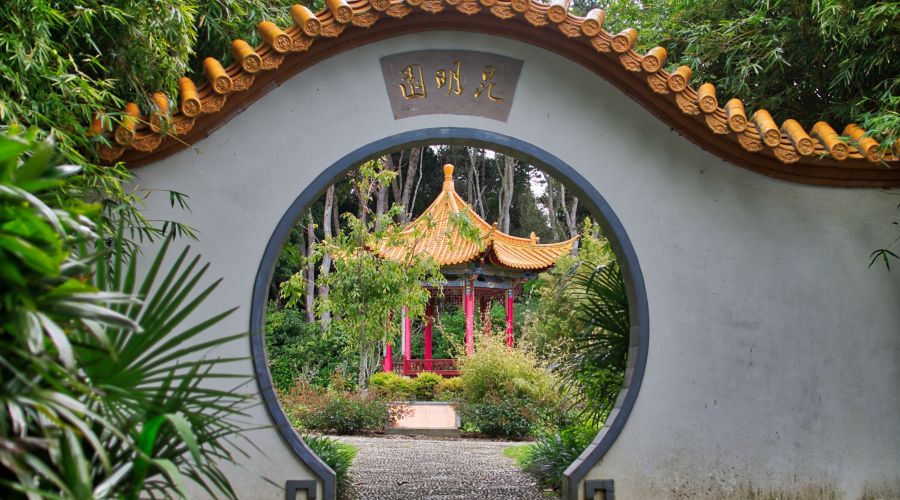 Ein traditionelles chinesisches Mondtor, eine runde Öffnung in einer weißen Gartenmauer, gibt den Blick frei auf einen Pagoden-Pavillon im Hintergrund. Üppige grüne Pflanzen und Bambus umrahmen die friedliche Szene.