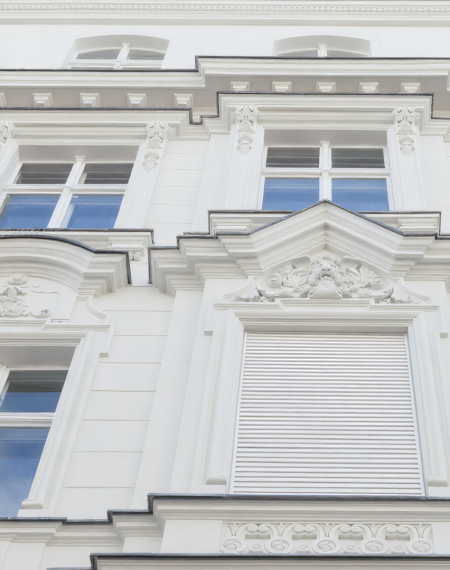 Die weiße Stuck-Fassade eines Altbaus wird von unten nach oben gezeigt. Mehrere verzierte Fenster sind zu sehen, eines davon ist mit einem geschlossenen Rollladen versehen, wie es für ältere Gebäude mit ungedämmten Kästen typisch ist.