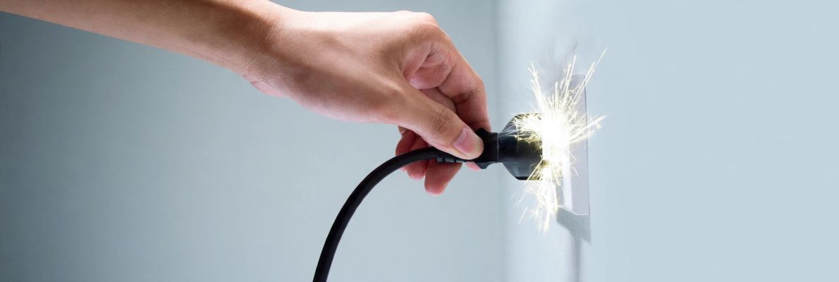 Eine Hand steckt ein schwarzes Stromkabel in eine weiße Wandsteckdose. Dabei entsteht ein heller, sprühender Funkenflug direkt am Kontakt zwischen dem Stecker und der Dose. Die Wand im Hintergrund ist schlicht und in einem hellen Grauton gehalten.