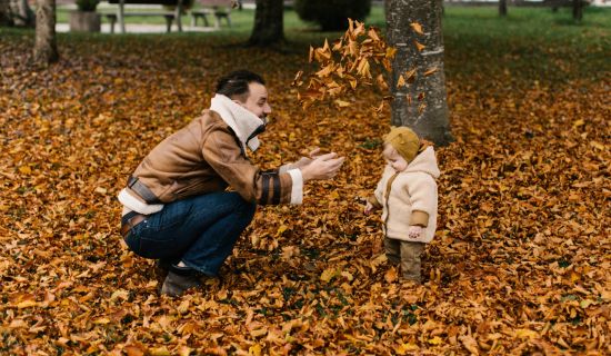 Ein Mann in einer braunen Jacke kniet im Freien auf einem Boden, der vollständig mit goldenen Herbstblättern bedeckt ist. Er spielt mit einem kleinen Kind in einer beigen Jacke, das vor ihm steht, und wirft Blätter in die Luft.