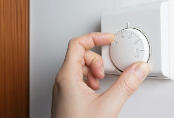 Eine Hand dreht einen Thermostatregler an einer Wand. Der Regler zeigt eine Temperaturanzeige, die auf etwa 20 Grad Celsius eingestellt ist.