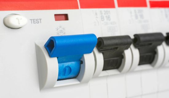 Nahaufnahme eines geöffneten Sicherungskastens mit Fokus auf einen blauen Fehlerstrom-Schutzschalter. Daneben befindet sich die weiße Test-Taste, während rechts weitere schwarze Leitungsschutzschalter aufgereiht sind.