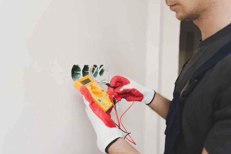 Elektriker mit rot-weißen Handschuhen misst mit einem gelben Multimeter die Spannung an Wandkabeln, die aus vier Öffnungen in der Wand ragen.
