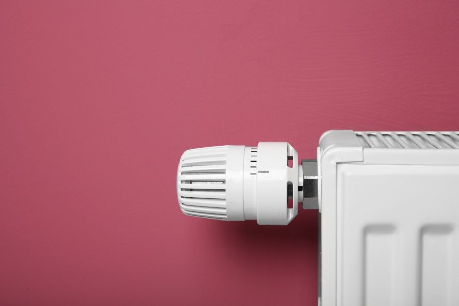 Ein weißer Heizkörperthermostat an einer Heizung vor einer einfarbigen, rötlich-pinken Wand.