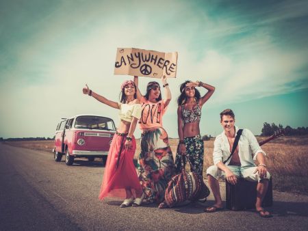 Vier junge Menschen in farbenfroher, gemusterter Kleidung stehen lachend an einer Landstraße neben einem rosa-weißen VW Bulli. Einer hält ein Pappschild mit der Aufschrift „Anywhere“ und einem Peace-Zeichen hoch.