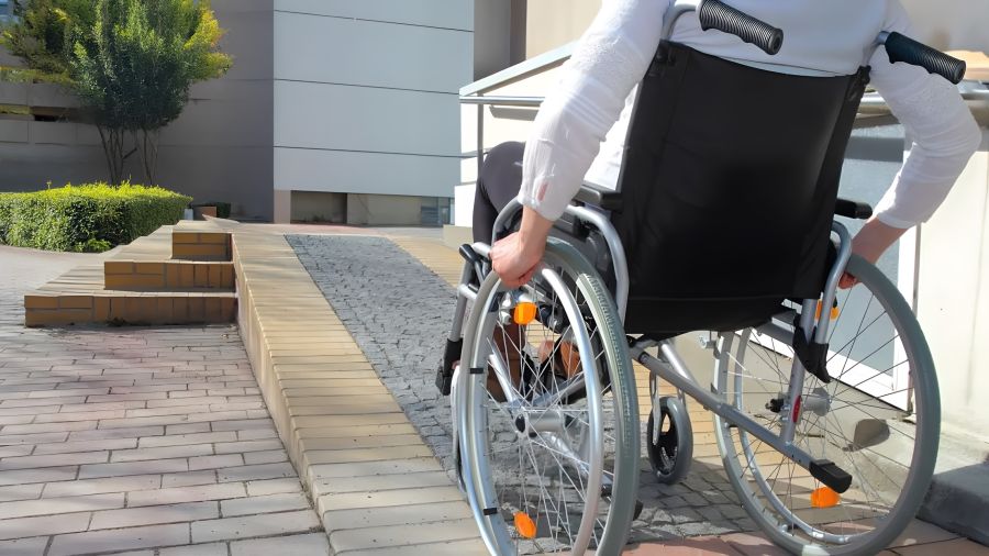Eine Person in einem Rollstuhl fährt eine gepflasterte Rampe hinauf. Die Umgebung besteht aus modernen Gebäuden und grünen Pflanzen.